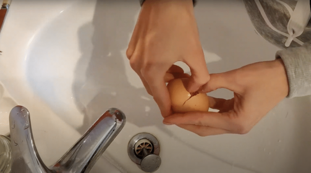 Two hands open a cracked egg over a sink, seen from above.