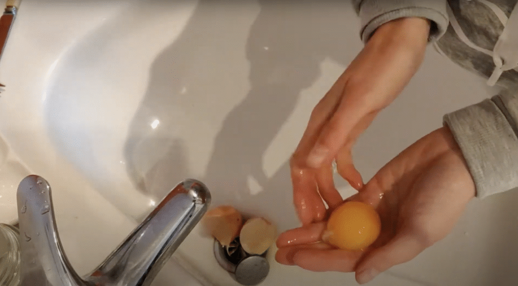 Seen from above, a pair of hands pass an egg yolk from side to side over a sink, an empty egg shell sits in the sink.