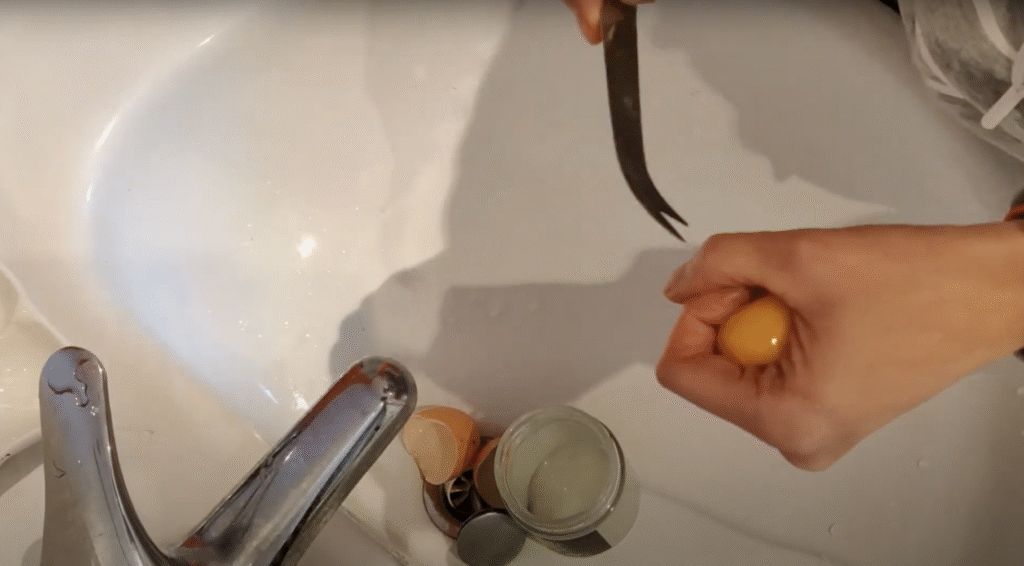 Seen from above, a hand holds an egg yolk gently in a fist, the other hand piercing the egg yolk membrane with the tip of a knife over a sink. A glass jar and empty egg shell sits in the sink.