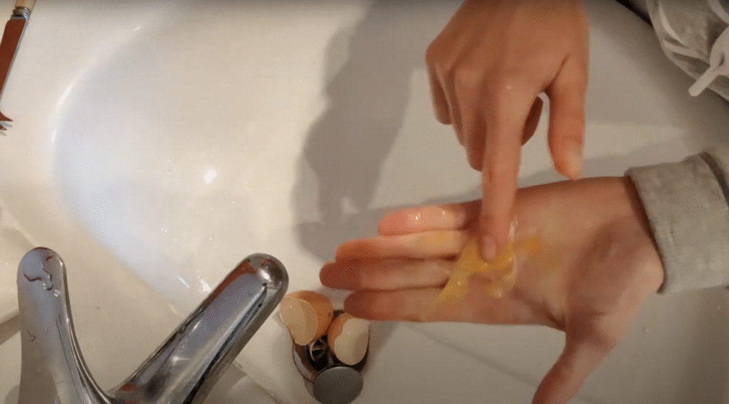 Seen from above, an open hand has an empty egg membrane sack in the palm, the other hand points to the membrane. The hands are over a sink with an empty egg shell in the sink.