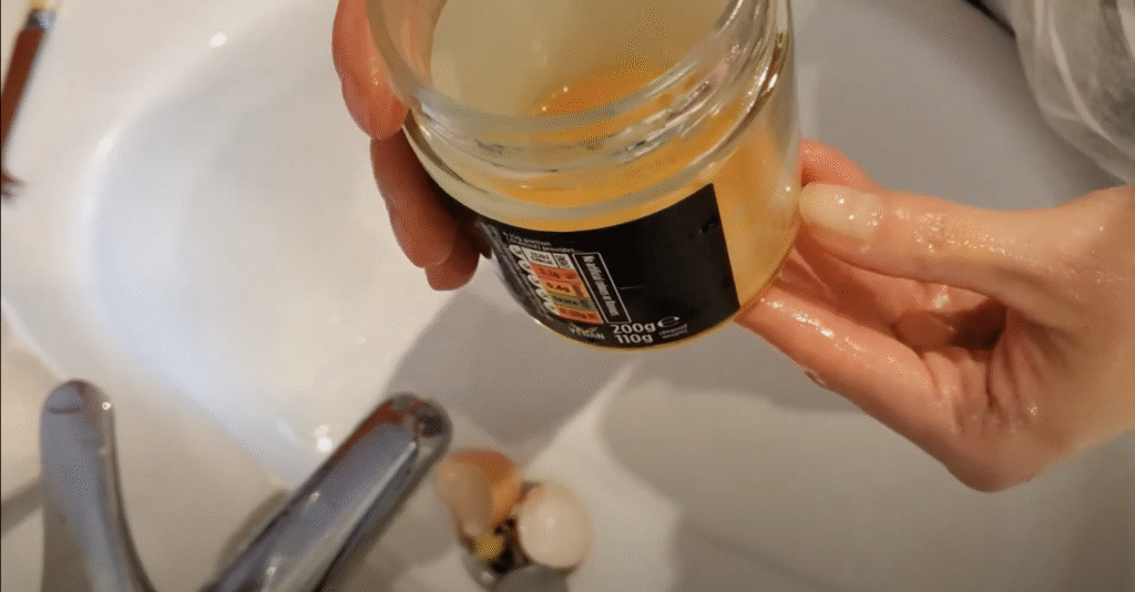 Seen from above, two hands hold a glass jar with egg yolk in over a sink, measuring how much egg yolk is in the jar using their thumb.