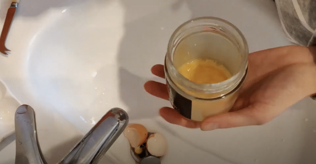 Seen from above, a hand holds a glass jar with egg yolk and white wine inside over a sink. The yolk has a frothy appearance and is the base for egg tempera paint.