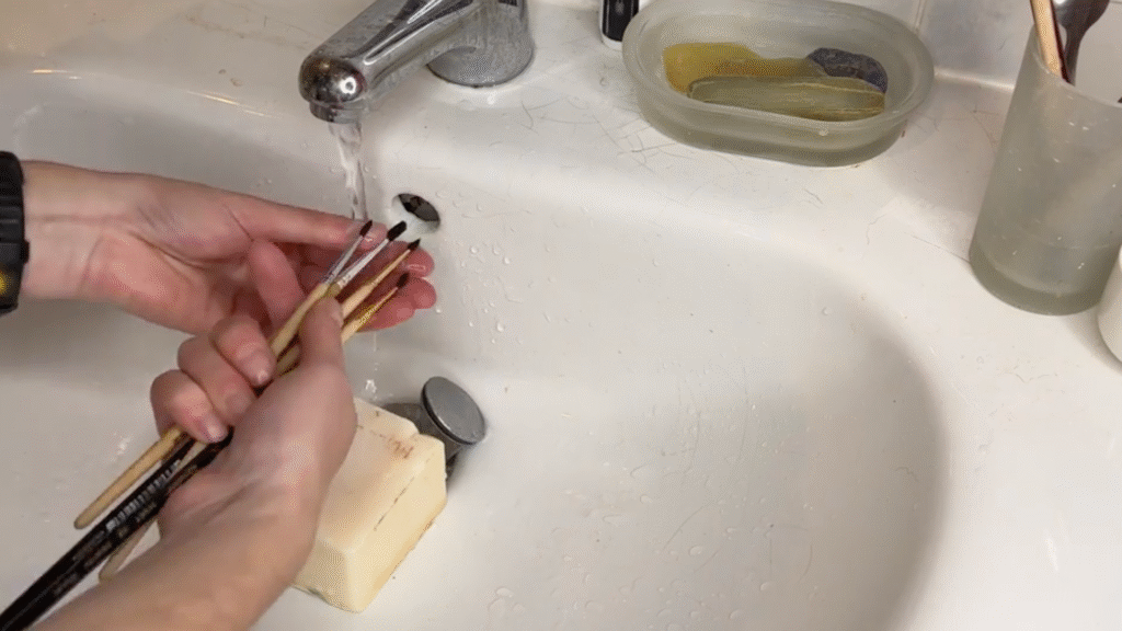 A tutorial for paintbrush care, a pair of hands rinse brushes under running water in a sink.