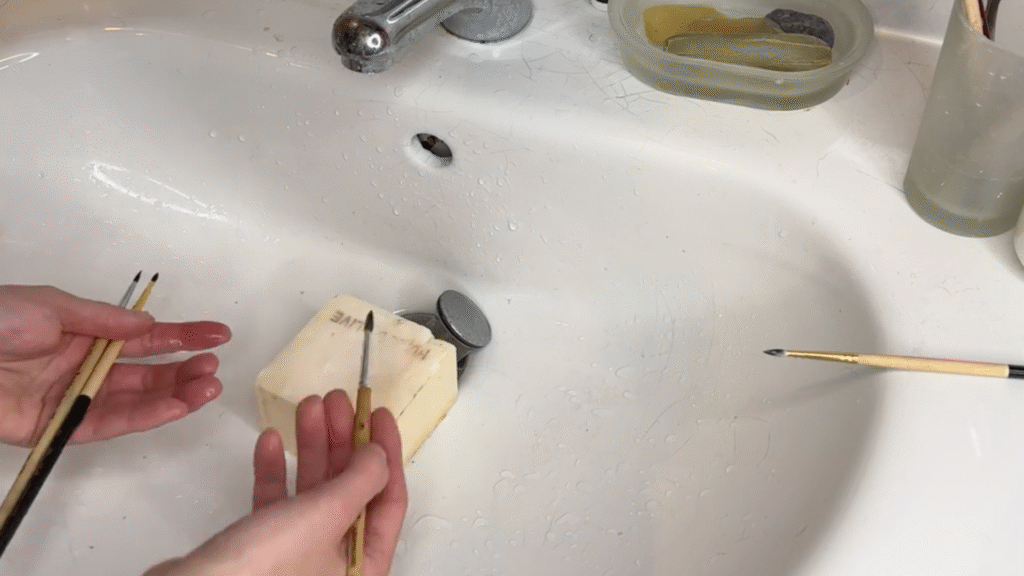 A tutorial for paintbrush care, a pair of hands rubs brushes onto soap in a sink.