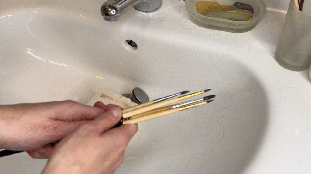 A tutorial for paintbrush care, a pair of hands hold soapy brushes in a sink.