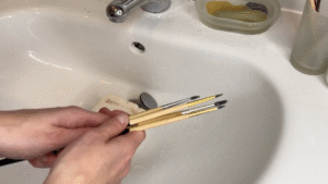 A tutorial for paintbrush care, a pair of hands hold soapy brushes in a sink.
