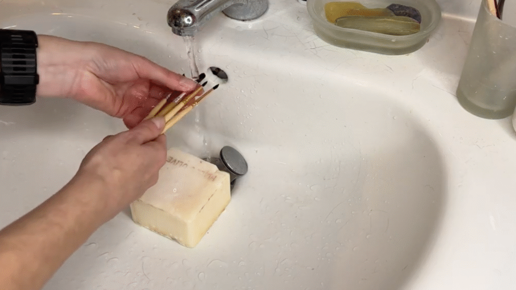 A tutorial for paintbrush care, a pair of hands rinse brushes under running water in a sink.